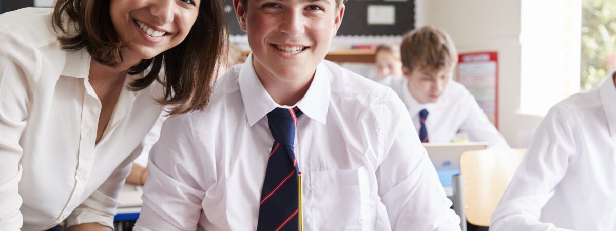Portrait Of Teacher Helping Pupil Using Computer In Classroom