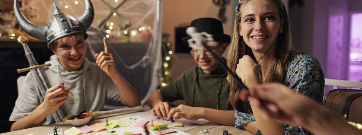 Three smiling teenagers in Halloween costumes play a board game together.