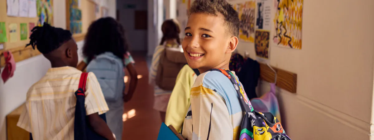 A smiling boy with a colorful backpack and blue folder looks back in a school hallway.