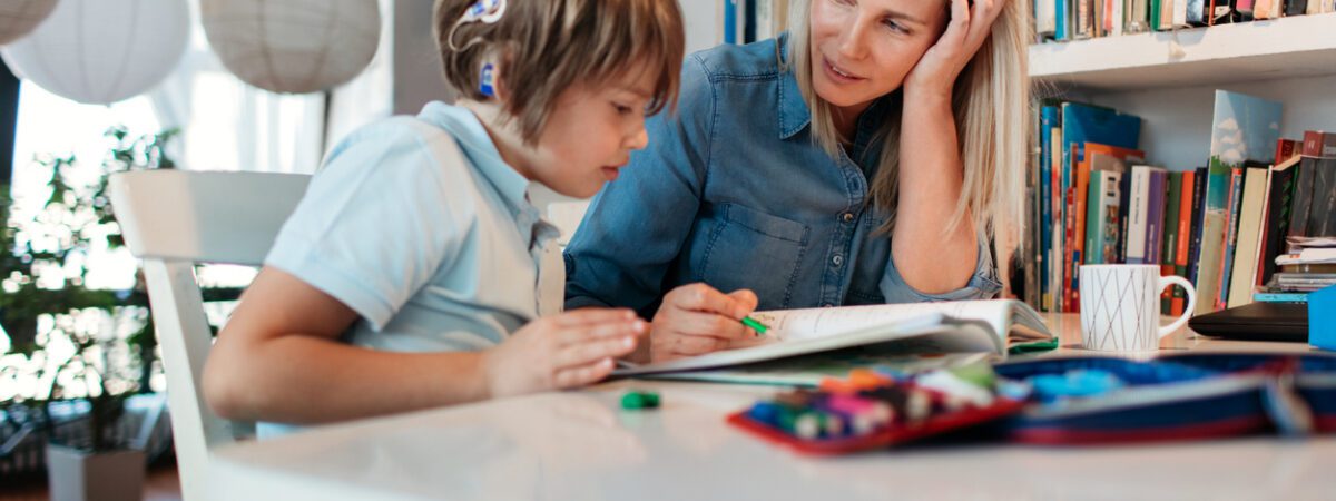 A mother helps her son, who wears hearing aids, with his homework at a table.