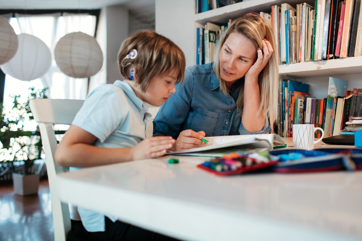 A mother helps her son, who wears hearing aids, with his homework at a table.