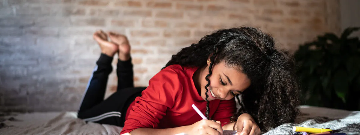 A smiling young girl with curly hair lies on a bed, writing in a notebook with a sandwich