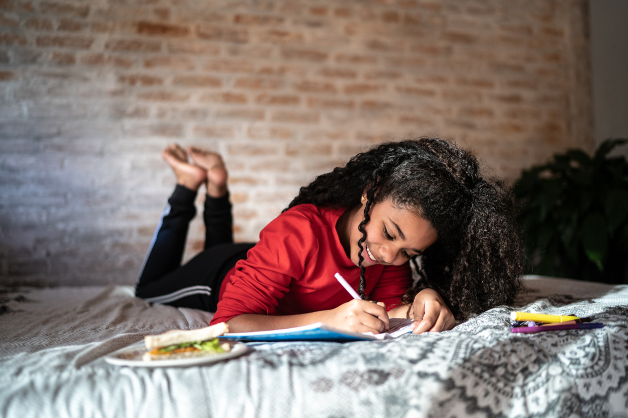 A smiling young girl with curly hair lies on a bed, writing in a notebook with a sandwich