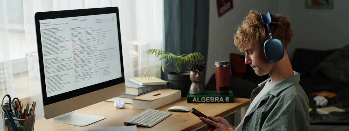 A young person wearing headphones scrolls on their phone at a desk with a computer displaying a school