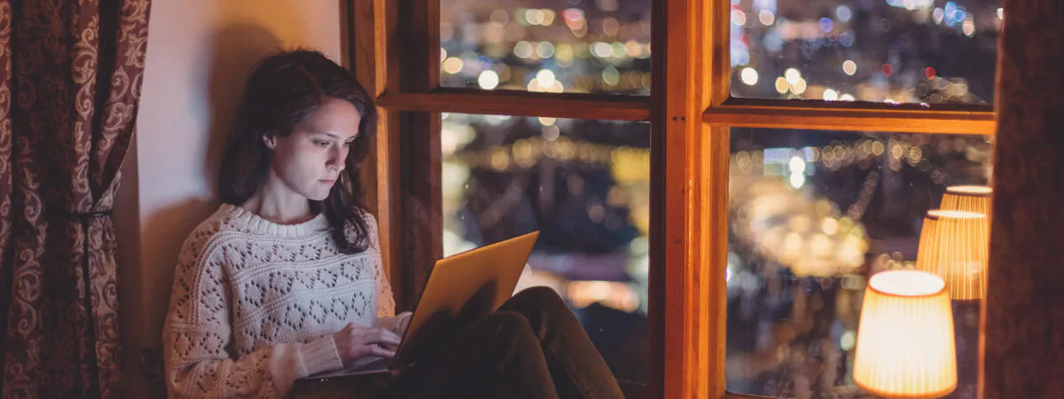 A young woman in a cozy sweater works on her laptop by a window overlooking city lights at