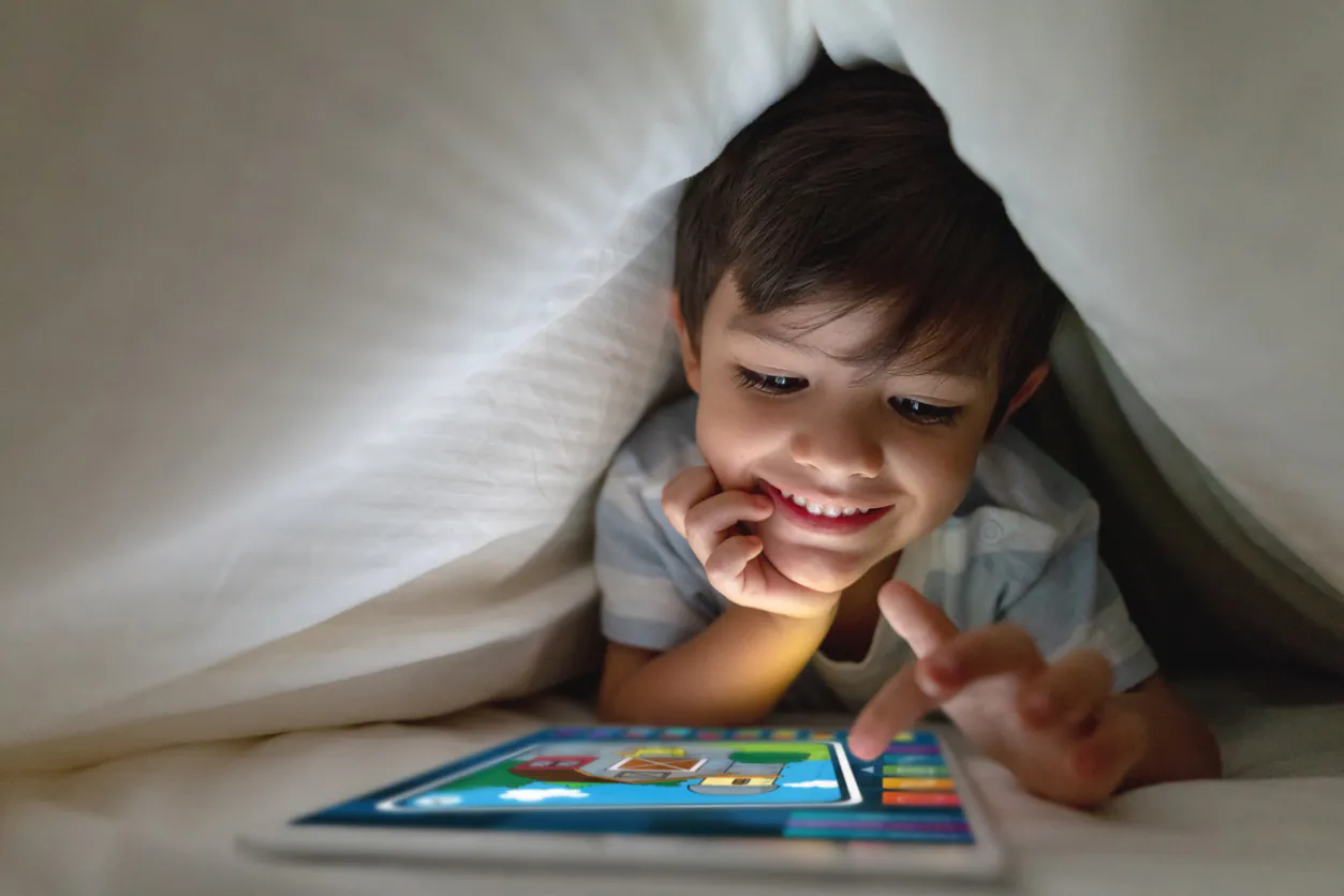 A smiling young boy plays a colorful game on a glowing tablet while tucked under white blankets.