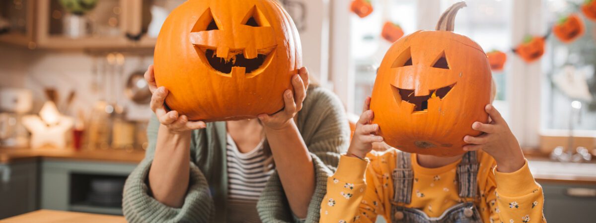 An adult and child hold carved jack-o'-lanterns over their faces in a festive
