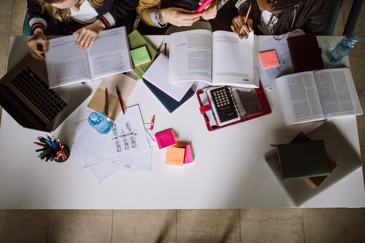 Students collaborate at a desk covered in open books, a laptop, and various study supplies.
